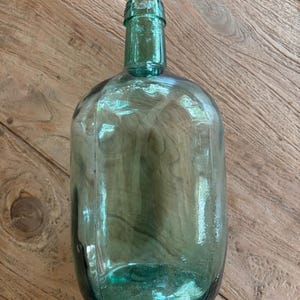 Puede incluir: Una botella de vidrio transl&uacute;cido en color verde mar con cuerpo redondeado y cuello estrecho. El vidrio presenta peque&ntilde;as burbujas de aire y una textura ligeramente irregular, con letras en relieve visibles en la parte superior del cuello.