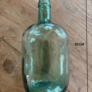 Puede incluir: Una botella de vidrio verde transl&uacute;cido con cuerpo redondeado y cuello estrecho. La botella presenta un texto en relieve claro en el borde superior. Las dimensiones indicadas por flechas muestran un ancho de 15 cm y una altura de 30 cm.