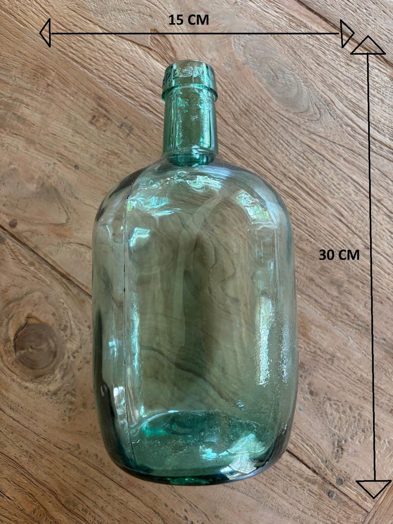 Puede incluir: Una botella de vidrio verde transl&uacute;cido con cuerpo redondeado y cuello estrecho. La botella presenta un texto en relieve claro en el borde superior. Las dimensiones indicadas por flechas muestran un ancho de 15 cm y una altura de 30 cm.
