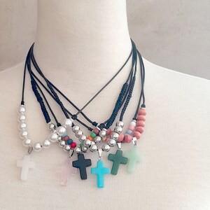 Peut inclure: Une collection de colliers superposés composés de cordons noirs et de chaînes argentées. Chaque collier comprend diverses perles blanches, roses, bleues, vertes et argentées, terminées par un pendentif en forme de croix de différentes couleurs.