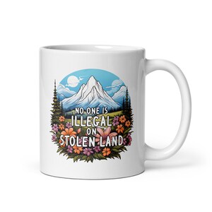 Könnte beinhalten: Weiße Keramiktasse mit einem Bergmotiv und dem Text "NO ONE IS ILLEGAL ON STOLEN LAND". Das Design ist von Blumen und Bäumen umgeben. Die Tasse hat einen gebogenen Griff.
