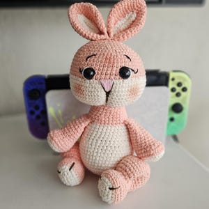 May include: A crocheted bunny toy in shades of pink and white. The bunny features large black eyes, a pink nose, and long ears. It is posed with its legs crossed. A video game console is visible in the background.