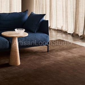 May include: A dark brown rectangular area rug with a smooth, uniform texture. The rug is positioned on a floor in front of a navy blue sofa, a light-colored pedestal side table, and a clear glass bowl. The text ArtisanLoomStudio is visible.