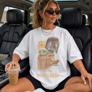 May include: A white short-sleeved t-shirt featuring a graphic print of the character Grogu and a Mandalorian helmet. The design includes a yellow sun, a small spacecraft, and the text "STAR WARS THE MANDALORIAN" printed in yellow lettering.