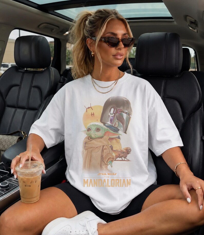 May include: A white short-sleeved t-shirt featuring a graphic print of the character Grogu and a Mandalorian helmet. The design includes a yellow sun, a small spacecraft, and the text "STAR WARS THE MANDALORIAN" printed in yellow lettering.