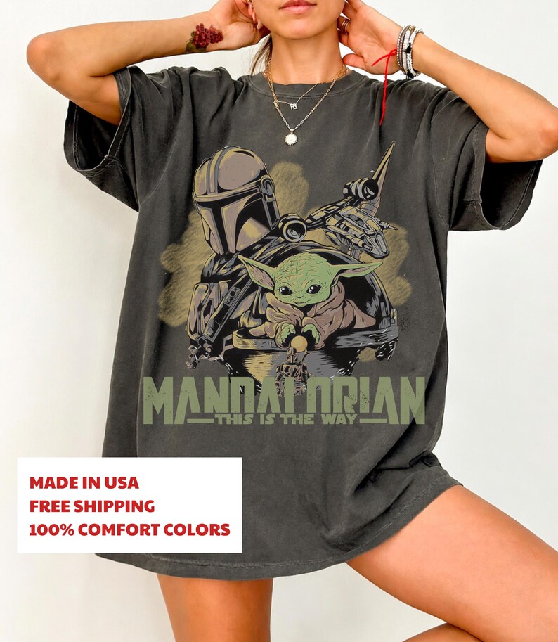May include: A charcoal gray short-sleeve t-shirt featuring a graphic print of a helmeted character and a small green creature. Below the illustration, the text reads MANDALORIAN THIS IS THE WAY in a muted olive green font with a distressed finish.