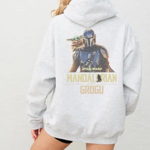 May include: A light gray hooded sweatshirt featuring a graphic print on the back of The Mandalorian and Grogu. The text reads Star Wars The Mandalorian and Grogu in a tan font. The hoodie has long sleeves and a hood with a drawstring closure.