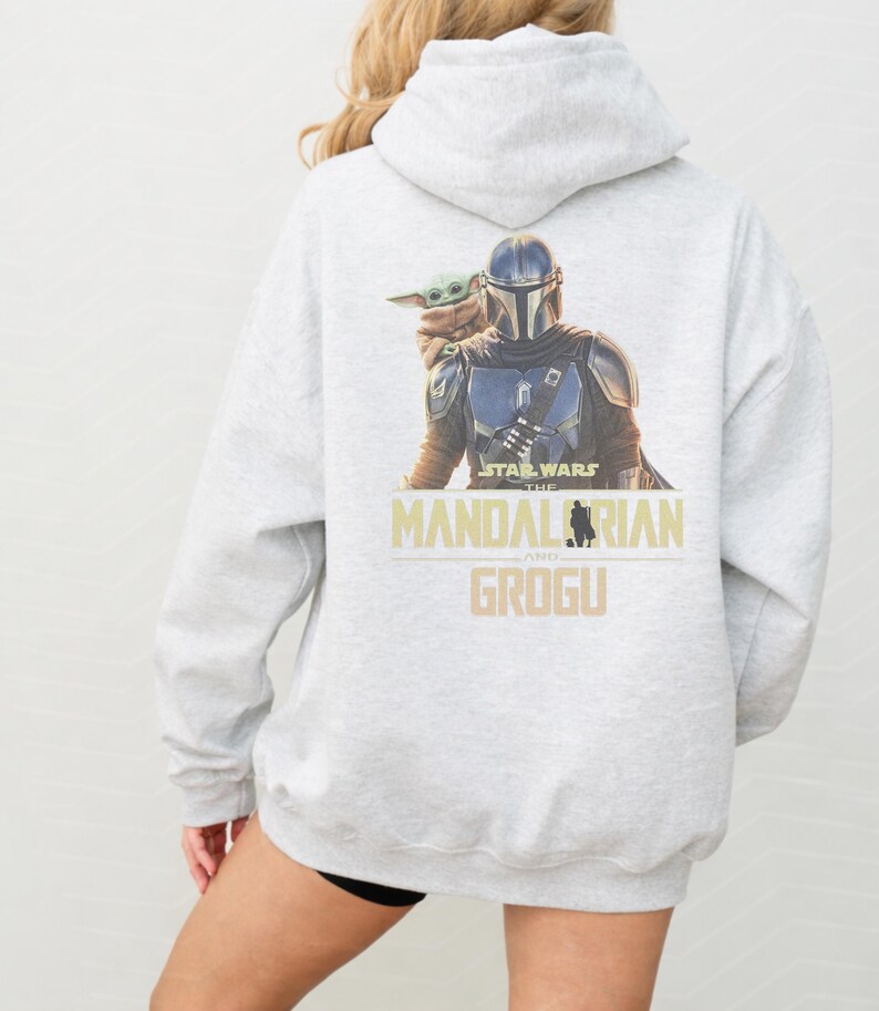 May include: A light gray hooded sweatshirt featuring a graphic print on the back of The Mandalorian and Grogu. The text reads Star Wars The Mandalorian and Grogu in a tan font. The hoodie has long sleeves and a hood with a drawstring closure.
