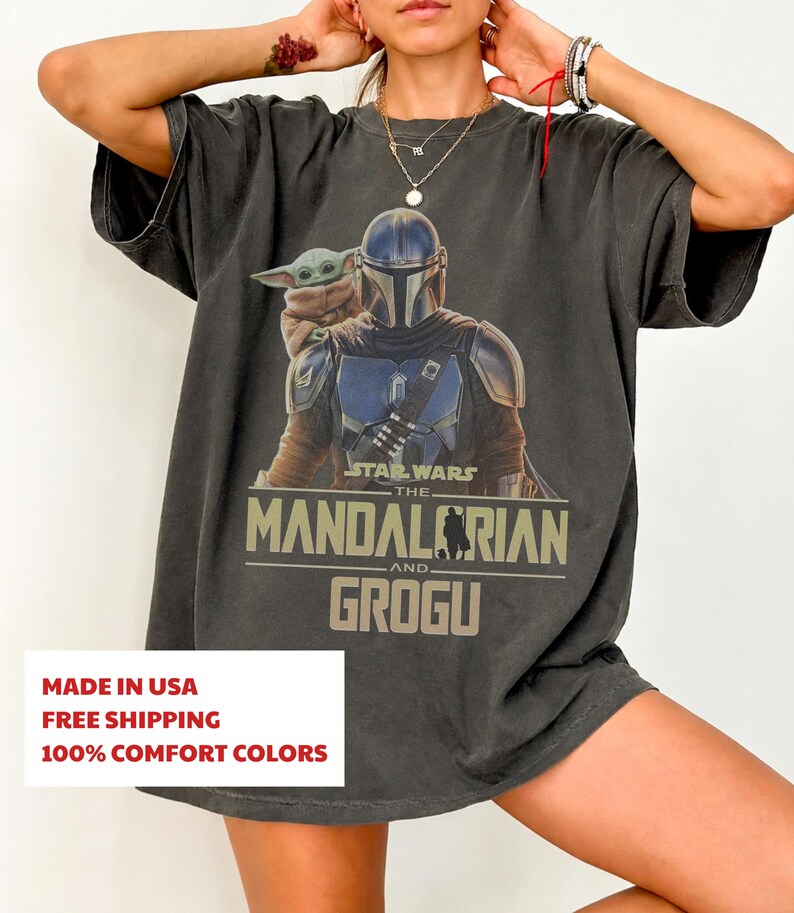 May include: A charcoal grey short-sleeve t-shirt featuring a graphic print of The Mandalorian and Grogu. The design includes the text Star Wars The Mandalorian and Grogu in a tan font. The shirt has a crew neckline and a relaxed, loose fit.