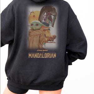May include: A black hooded sweatshirt featuring a graphic print on the back. The design displays a green creature with large ears, a metallic helmet, a yellow sun, and a small spacecraft. The text reads STAR WARS THE MANDALORIAN in gold letters.