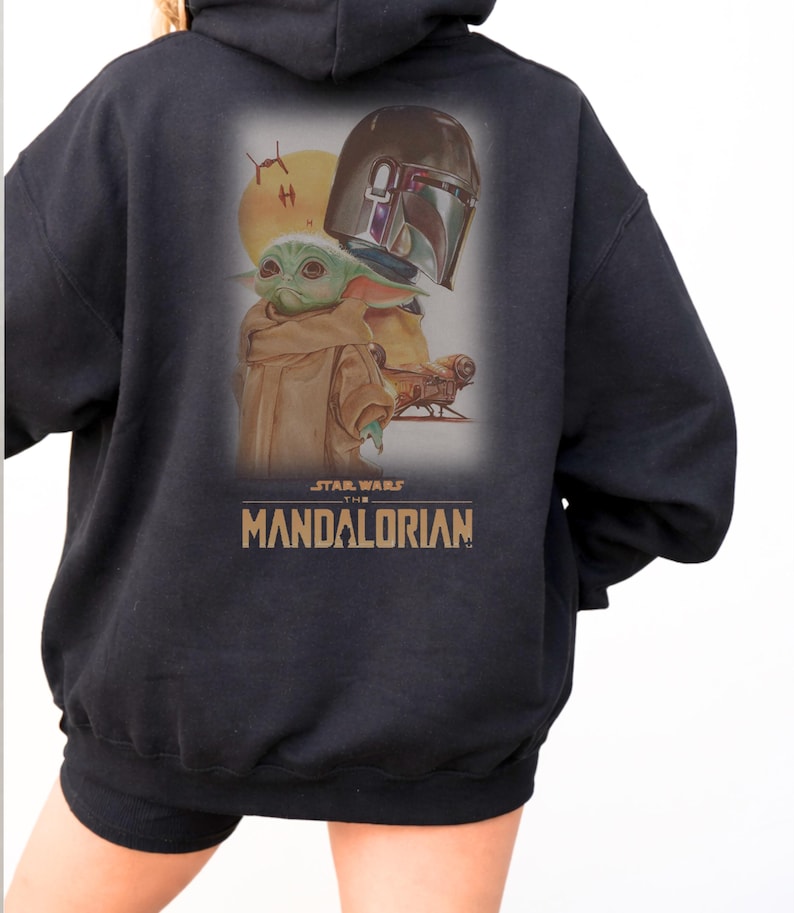 May include: A black hooded sweatshirt featuring a graphic print on the back. The design displays a green creature with large ears, a metallic helmet, a yellow sun, and a small spacecraft. The text reads STAR WARS THE MANDALORIAN in gold letters.
