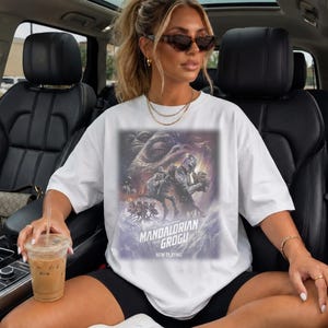 May include: A white short-sleeved t-shirt featuring a graphic print of The Mandalorian and Grogu. The design includes the text Star Wars The Mandalorian and Grogu Now Playing, showcasing characters from the series in a cinematic poster style.