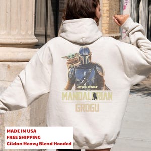 May include: An off-white hooded sweatshirt featuring a graphic print on the back. The design displays The Mandalorian in armor with Grogu on a shoulder. Below the characters, the text reads STAR WARS THE MANDALORIAN AND GROGU in yellow.