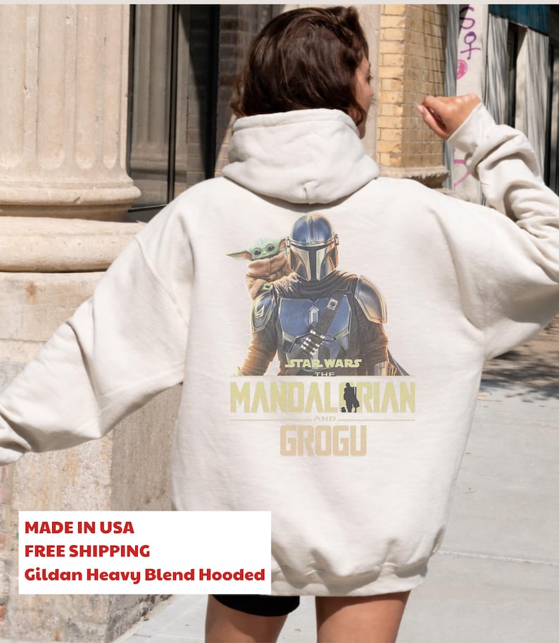 May include: An off-white hooded sweatshirt featuring a graphic print on the back. The design displays The Mandalorian in armor with Grogu on a shoulder. Below the characters, the text reads STAR WARS THE MANDALORIAN AND GROGU in yellow.