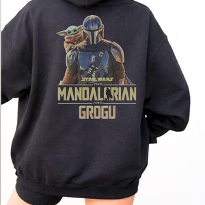 May include: A black hooded sweatshirt featuring a graphic print on the back. The design displays The Mandalorian and Grogu with the text Star Wars The Mandalorian and Grogu in tan lettering. The hoodie has long sleeves and a drawstring hood.