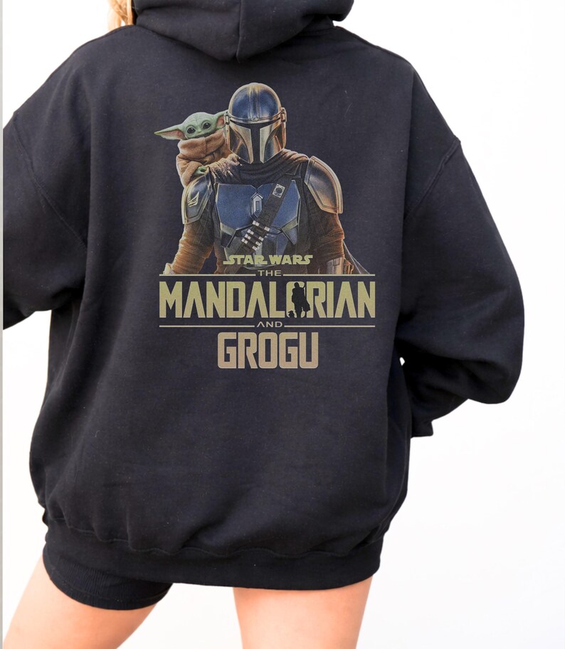 May include: A black hooded sweatshirt featuring a graphic print on the back. The design displays The Mandalorian and Grogu with the text Star Wars The Mandalorian and Grogu in tan lettering. The hoodie has long sleeves and a drawstring hood.