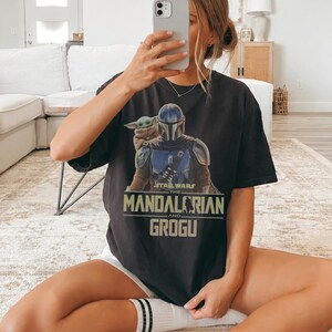 May include: A black short-sleeve t-shirt featuring a graphic print of The Mandalorian and Grogu. The design includes the text Star Wars The Mandalorian and Grogu in a light green font. The shirt has a crew neckline and a relaxed fit.