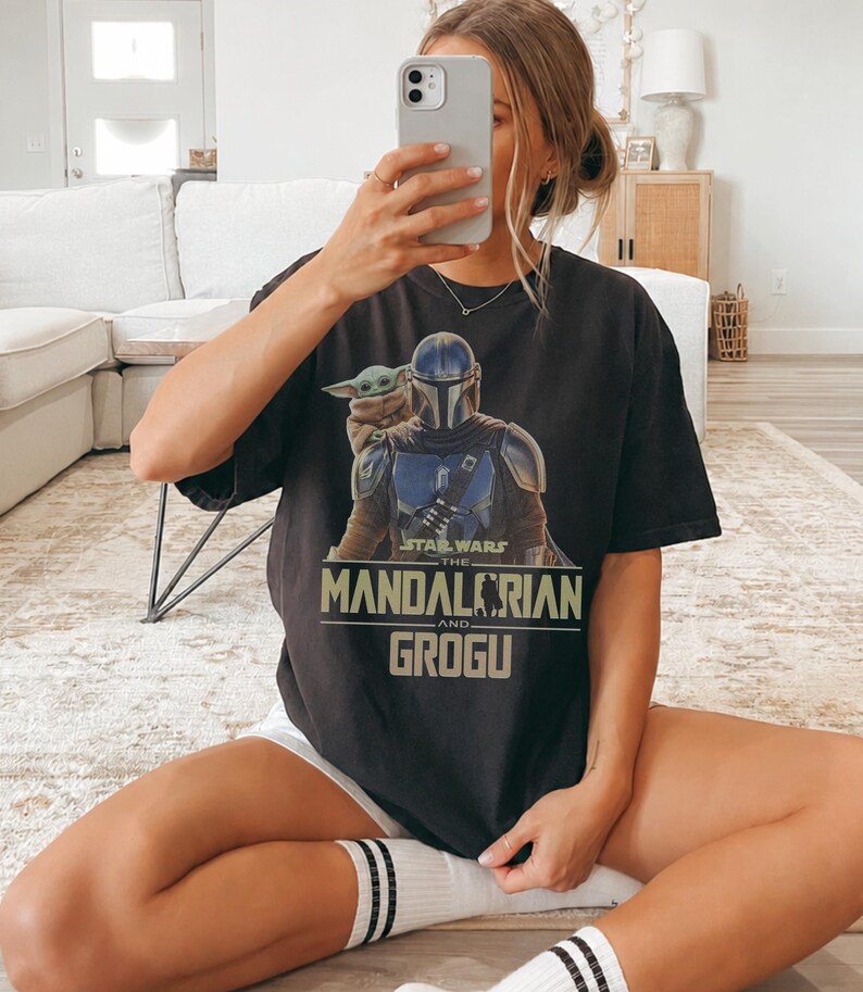 May include: A black short-sleeve t-shirt featuring a graphic print of The Mandalorian and Grogu. The design includes the text Star Wars The Mandalorian and Grogu in a light green font. The shirt has a crew neckline and a relaxed fit.