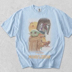 May include: A light blue t-shirt featuring a graphic print of The Mandalorian and Grogu. The design includes a yellow sun, two small starships, and the text Star Wars The Mandalorian in gold lettering. The shirt has a crew neck and short sleeves.
