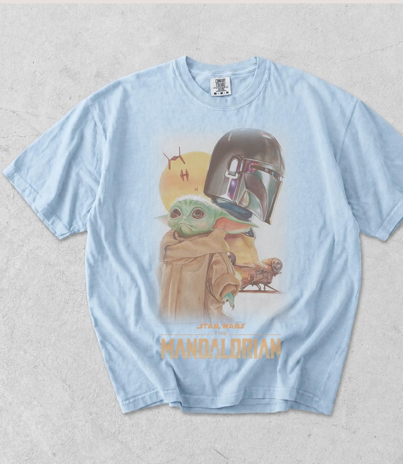 May include: A light blue t-shirt featuring a graphic print of The Mandalorian and Grogu. The design includes a yellow sun, two small starships, and the text Star Wars The Mandalorian in gold lettering. The shirt has a crew neck and short sleeves.
