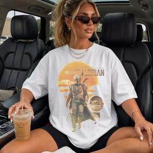 K&ouml;nnte beinhalten: Ein wei&szlig;es Kurzarm-T-Shirt mit einem Grafikdruck der Figur The Mandalorian und Grogu in einer schwebenden Kapsel vor einem gelben Sonnenhintergrund. Der Text THE MANDALORIAN ist in fetter Schrift &uuml;ber der zentralen Illustration aufgedruckt.