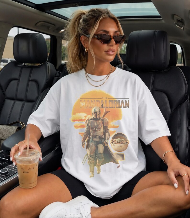 K&ouml;nnte beinhalten: Ein wei&szlig;es Kurzarm-T-Shirt mit einem Grafikdruck der Figur The Mandalorian und Grogu in einer schwebenden Kapsel vor einem gelben Sonnenhintergrund. Der Text THE MANDALORIAN ist in fetter Schrift &uuml;ber der zentralen Illustration aufgedruckt.