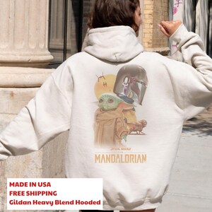 May include: A cream-colored hooded sweatshirt featuring a graphic on the back with a small green creature, a metallic helmet, and a spaceship. The text "STAR WARS THE MANDALORIAN" is printed in orange below the illustration on the back of the garment.