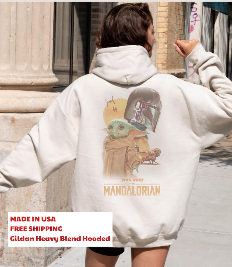 May include: A cream-colored hooded sweatshirt featuring a graphic on the back with a small green creature, a metallic helmet, and a spaceship. The text "STAR WARS THE MANDALORIAN" is printed in orange below the illustration on the back of the garment.