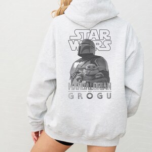 Może przedstawiać: Jasnoszara bluza z kapturem z czarno-białą grafiką na plecach. Wz&oacute;r przedstawia logo Star Wars nad ilustracją Mandalorianina trzymającego Grogu, z napisem Mandalorian and Grogu wydrukowanym poniżej.