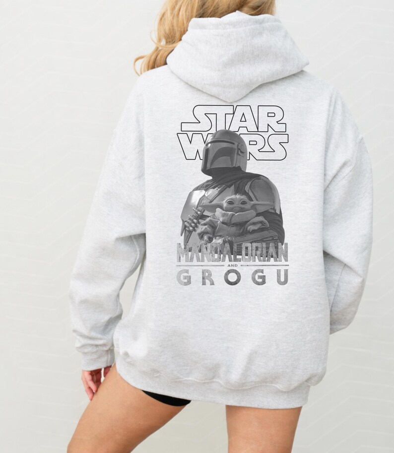 Może przedstawiać: Jasnoszara bluza z kapturem z czarno-białą grafiką na plecach. Wz&oacute;r przedstawia logo Star Wars nad ilustracją Mandalorianina trzymającego Grogu, z napisem Mandalorian and Grogu wydrukowanym poniżej.