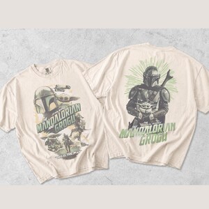 Puede incluir: Una camiseta de color crema con gr&aacute;ficos de The Mandalorian y Grogu en la parte delantera y trasera. El frente muestra varias escenas con el texto The Mandalorian and Grogu y This Is The Way. La parte posterior muestra una figura mandaloriana.