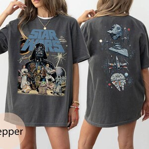May include: A charcoal gray short-sleeve t-shirt featuring a Star Wars graphic. The front displays Darth Vader, Luke Skywalker, Princess Leia, Chewbacca, and other characters. The back shows a vertical arrangement of the Death Star and starships.