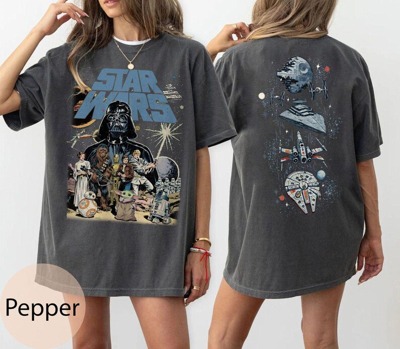May include: A charcoal gray short-sleeve t-shirt featuring a Star Wars graphic. The front displays Darth Vader, Luke Skywalker, Princess Leia, Chewbacca, and other characters. The back shows a vertical arrangement of the Death Star and starships.