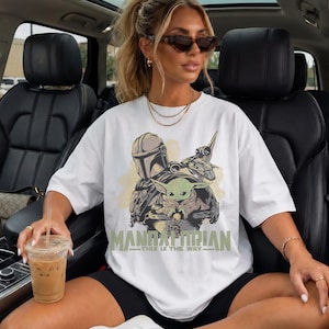 May include: A white short-sleeved t-shirt featuring a graphic print of a helmeted character and a small green creature in a pod. The text "MANDALORIAN" and "THIS IS THE WAY" is printed in a muted green font below the central character illustration.