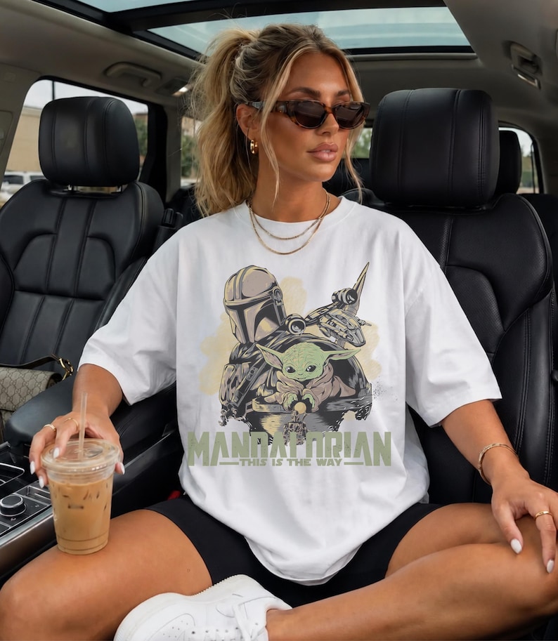 May include: A white short-sleeved t-shirt featuring a graphic print of a helmeted character and a small green creature in a pod. The text "MANDALORIAN" and "THIS IS THE WAY" is printed in a muted green font below the central character illustration.
