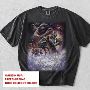 May include: A charcoal grey short-sleeve t-shirt featuring a graphic print of The Mandalorian and Grogu. The design includes a large eye, space scenes, and the text THE MANDALORIAN AND GROGU NOW PLAYING. The shirt has a crew neckline and a relaxed fit.