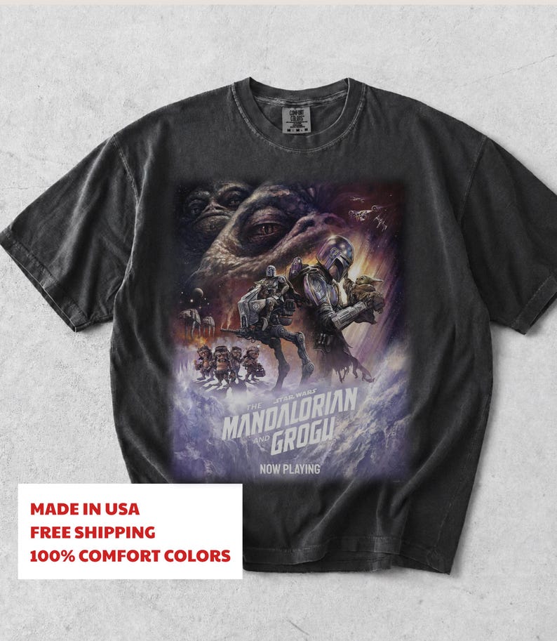 May include: A charcoal grey short-sleeve t-shirt featuring a graphic print of The Mandalorian and Grogu. The design includes a large eye, space scenes, and the text THE MANDALORIAN AND GROGU NOW PLAYING. The shirt has a crew neckline and a relaxed fit.