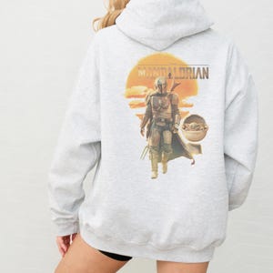 K&ouml;nnte beinhalten: Ein hellgrauer Kapuzenpullover mit einem Grafikdruck auf der R&uuml;ckseite. Das Motiv zeigt den Mandalorianer in R&uuml;stung, der vor einer gro&szlig;en orangefarbenen Sonne geht, daneben eine kleine Kreatur in einer schwebenden Kapsel und der Schriftzug The Mandalorian.