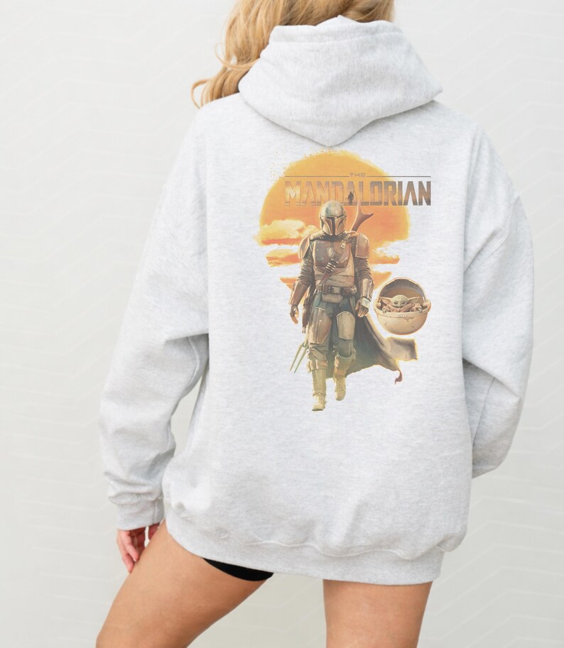 K&ouml;nnte beinhalten: Ein hellgrauer Kapuzenpullover mit einem Grafikdruck auf der R&uuml;ckseite. Das Motiv zeigt den Mandalorianer in R&uuml;stung, der vor einer gro&szlig;en orangefarbenen Sonne geht, daneben eine kleine Kreatur in einer schwebenden Kapsel und der Schriftzug The Mandalorian.