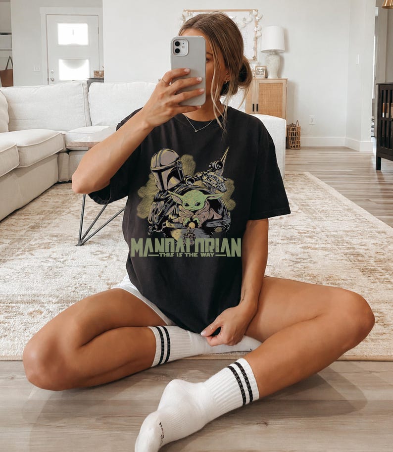 May include: A black short-sleeve t-shirt featuring a graphic print of The Mandalorian and Grogu. The design includes the text "MANDALORIAN" in large green letters and "THIS IS THE WAY" in smaller green text below the character illustration.
