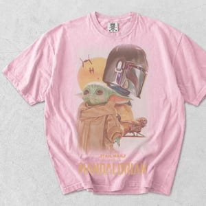 May include: A light pink short-sleeve t-shirt featuring a graphic print of Grogu and the Mandalorian helmet. The design includes a yellow sun, small starships, and the text Star Wars The Mandalorian in yellow lettering at the bottom center.