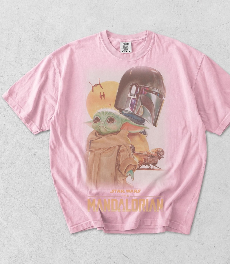 May include: A light pink short-sleeve t-shirt featuring a graphic print of Grogu and the Mandalorian helmet. The design includes a yellow sun, small starships, and the text Star Wars The Mandalorian in yellow lettering at the bottom center.