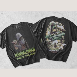 May include: A black short-sleeve t-shirt featuring a graphic of The Mandalorian and Grogu. The front displays the characters with the text "THE MANDALORIAN THIS IS THE WAY". The back features a larger collage of the characters and the text "THE MANDALORIAN AND GROGU" and "THIS IS THE WAY".