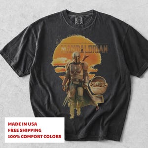 K&ouml;nnte beinhalten: Ein anthrazitfarbenes Kurzarm-T-Shirt mit einem Grafikdruck des Mandalorianers und Grogu in einer schwebenden Kapsel. Das Design zeigt einen gro&szlig;en orangefarbenen Sonnenhintergrund und den Text THE MANDALORIAN in einer fetten, abgenutzten Schrift.