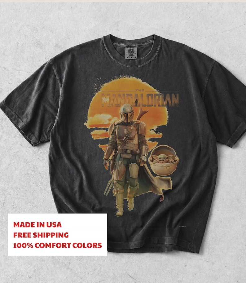 K&ouml;nnte beinhalten: Ein anthrazitfarbenes Kurzarm-T-Shirt mit einem Grafikdruck des Mandalorianers und Grogu in einer schwebenden Kapsel. Das Design zeigt einen gro&szlig;en orangefarbenen Sonnenhintergrund und den Text THE MANDALORIAN in einer fetten, abgenutzten Schrift.