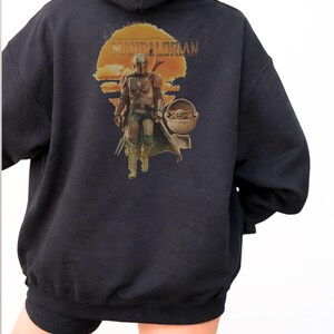 K&ouml;nnte beinhalten: Ein schwarzer Kapuzenpullover mit einem Aufdruck auf der R&uuml;ckseite. Das Motiv zeigt eine Figur in metallischer R&uuml;stung, die vor einer gro&szlig;en orangefarbenen Sonne geht, mit dem Text THE MANDALORIAN dar&uuml;ber. Neben der Figur ist ein kleines Wesen.