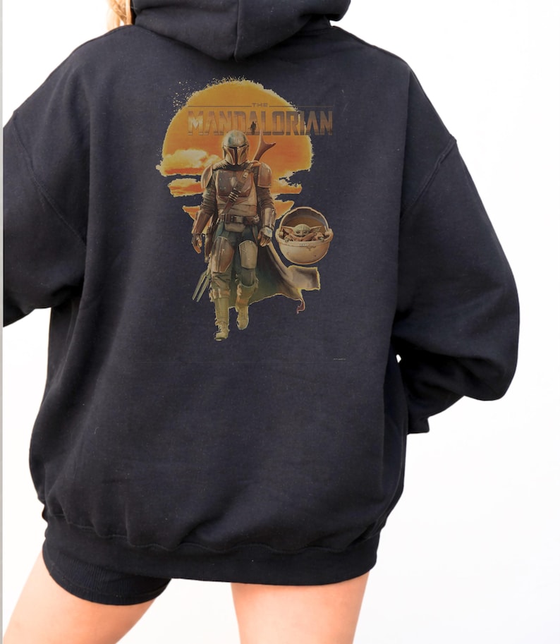 K&ouml;nnte beinhalten: Ein schwarzer Kapuzenpullover mit einem Aufdruck auf der R&uuml;ckseite. Das Motiv zeigt eine Figur in metallischer R&uuml;stung, die vor einer gro&szlig;en orangefarbenen Sonne geht, mit dem Text THE MANDALORIAN dar&uuml;ber. Neben der Figur ist ein kleines Wesen.