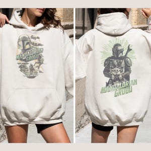 Puede incluir: Una sudadera con capucha de color crema con estampados gr&aacute;ficos de The Mandalorian y Grogu. El frente muestra un collage de personajes con el texto The Mandalorian and Grogu. La parte posterior muestra una gran ilustraci&oacute;n del d&uacute;o con rayos verdes.