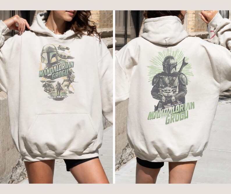 Puede incluir: Una sudadera con capucha de color crema con estampados gr&aacute;ficos de The Mandalorian y Grogu. El frente muestra un collage de personajes con el texto The Mandalorian and Grogu. La parte posterior muestra una gran ilustraci&oacute;n del d&uacute;o con rayos verdes.