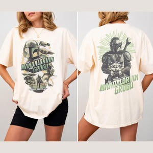 Puede incluir: Una camiseta de manga corta color hueso con gr&aacute;ficos de Star Wars. El frente muestra a The Mandalorian y Grogu con el texto "The Mandalorian and Grogu" y "This is the way". La parte posterior muestra una gran ilustraci&oacute;n de los personajes.